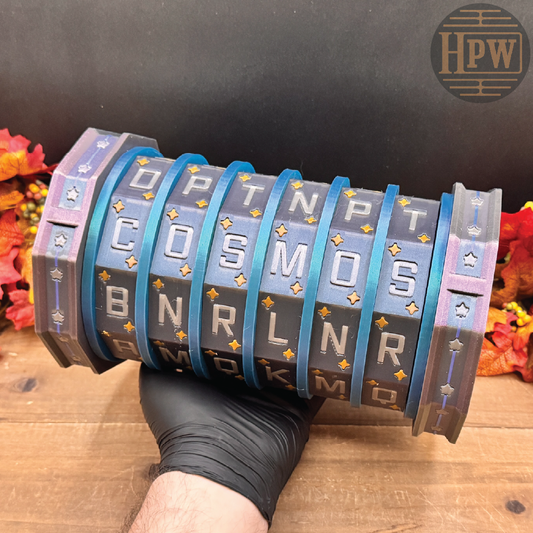 A hand wearing a black glove holds a cylindrical, multi-panel alphabet calculator toy. The panels are dark gray with silver lettering, accented by bright blue bands and gold star details.
