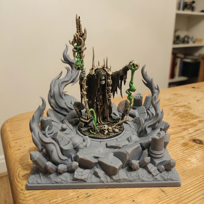 A painted fantasy tabletop miniature display featuring a dark robed figure holding staffs, surrounded by gray flames, rocks, and green vine details, set on a wooden table indoors.