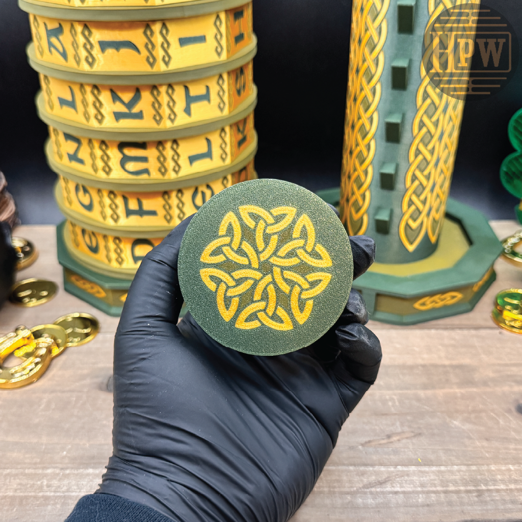 Hand holding a green and gold circular object with Celtic knot design, surrounded by similar objects on a wooden surface.