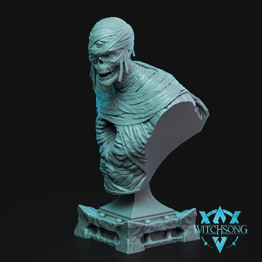 A 3D-printed resin figurine of a mummy bust on a display base, shown in a light teal-gray color against a solid black background. The Witchsong logo is visible in the bottom right corner.