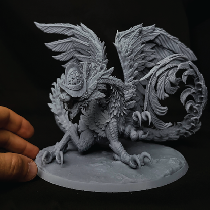 A detailed 3D-printed unpainted fantasy miniature of a griffin wearing a hat, shown with a human hand holding the base against a black background.