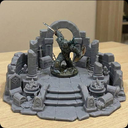 A tabletop miniature display featuring a fantasy creature figurine posed in a ruined stone arena with stairs, arches, crates, and debris, photographed on a light wood surface against a beige indoor background.