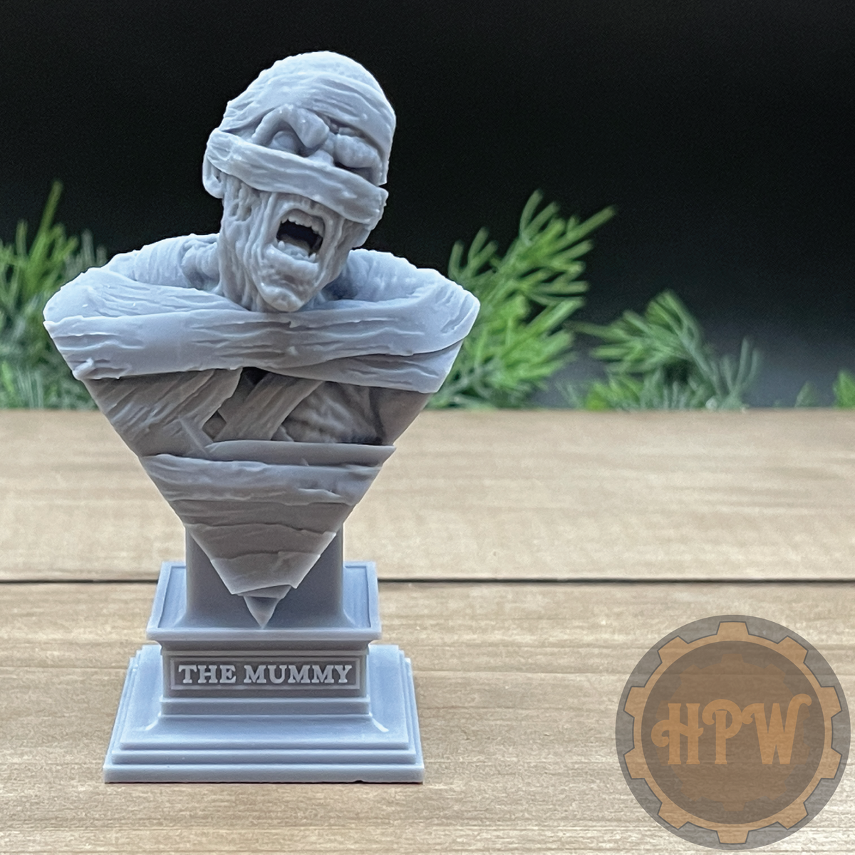 A gray 3D-printed bust sculpture of 'The Mummy' on a tiered display base, set on a wooden tabletop with a dark background and green foliage.