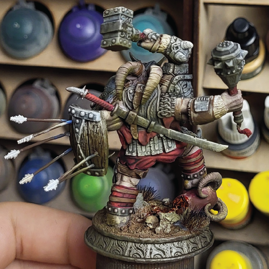 Detailed painted miniature of a demon goat warrior with armor and weapons on a dark background seen from the rear in front of a painting station
