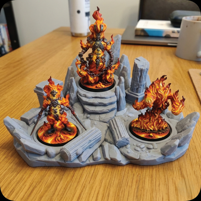 Three fantasy tabletop miniatures with glowing orange and yellow flames on a gray rocky display base, photographed on a wooden desk with a laptop and mug in the background.