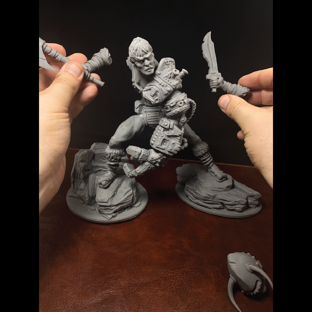 A gray unpainted fantasy warrior tabletop miniature is posed on a brown wooden surface against a dark background, with two hands holding additional unpainted accessories and a small gray item in the lower right.