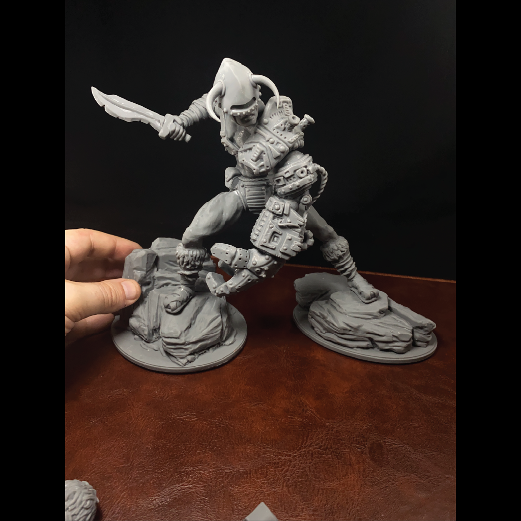 A gray unpainted 3D-printed fantasy miniature figurine of a warrior stepping over a fallen opponent, displayed on a brown leather surface against a black background, with a hand holding the left side of the base.