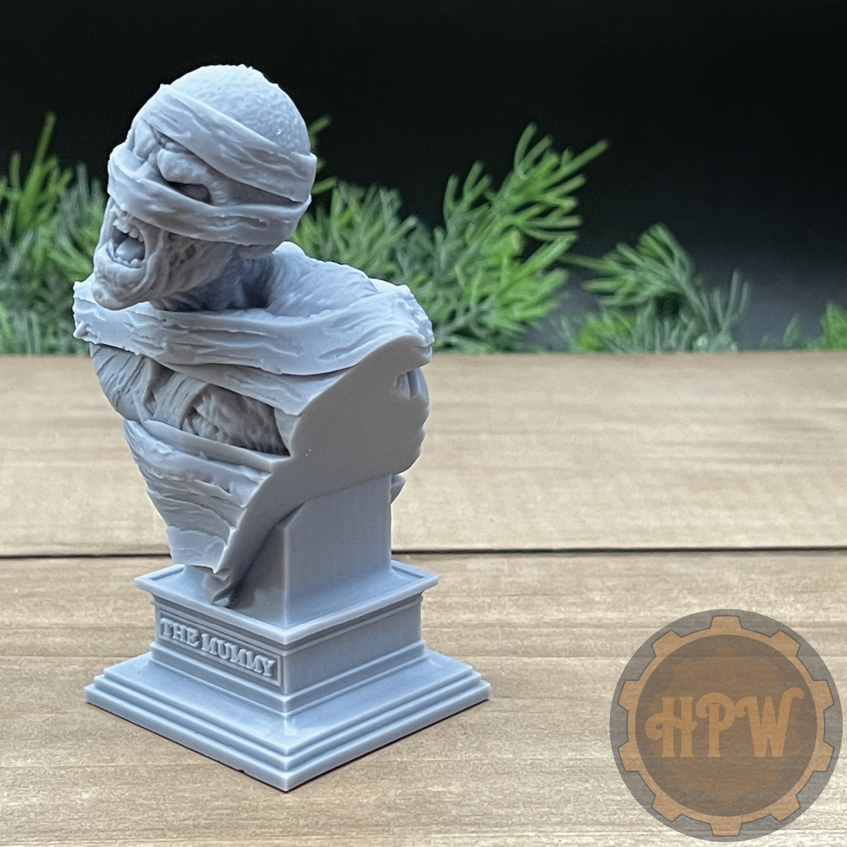 A gray 3D-printed bust of a mummy wrapped in bandages, mounted on a tiered pedestal base labeled 'THE MUMMY', set on a wooden tabletop with blurred green foliage in the background.
