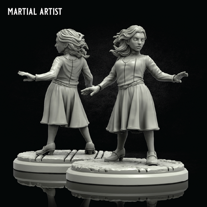 Two unpainted miniature figurines of women in combat poses on round bases, shown against a black background with the text 'MARTIAL ARTIST' in the top left corner.