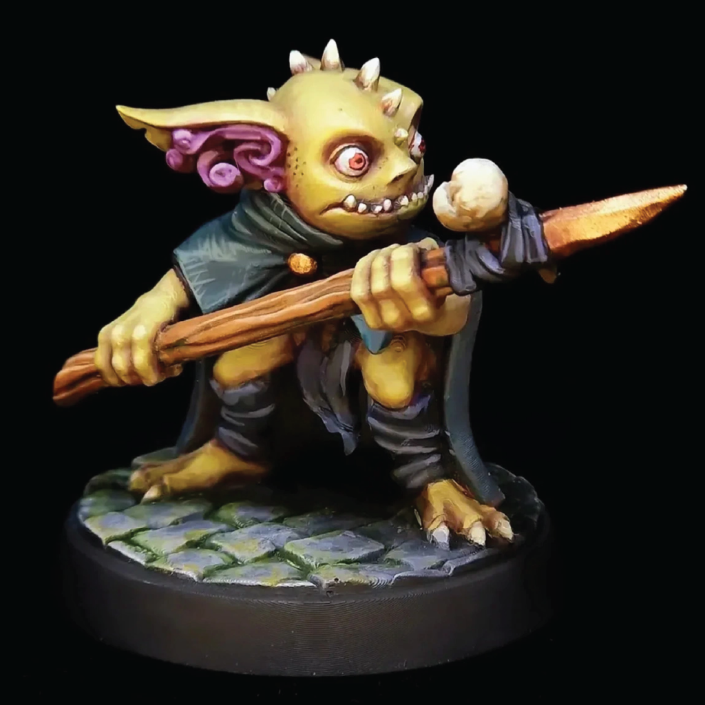 A painted fantasy tabletop miniature figurine of a small green-skinned creature with horns, purple ears, and a dark cloak, holding a wooden staff with a glowing orb. The figure is posed on a stone-textured base against a black background.