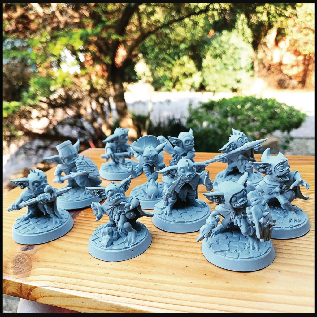 A group of unpainted gray fantasy tabletop gaming miniatures arranged on a wooden table outdoors, with a blurred garden background.