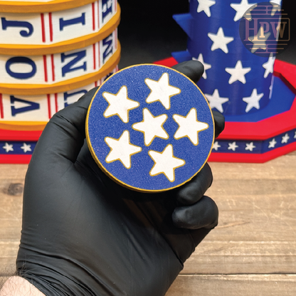 A hand wearing a black glove holds a round blue coaster with five white stars, with patriotic-themed coasters in the background on a wooden surface.