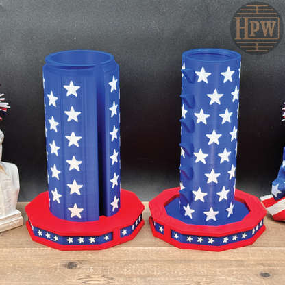 Two patriotic-themed cylindrical holders with blue bodies covered in white stars and red octagonal bases, displayed on a wooden surface against a dark backdrop.