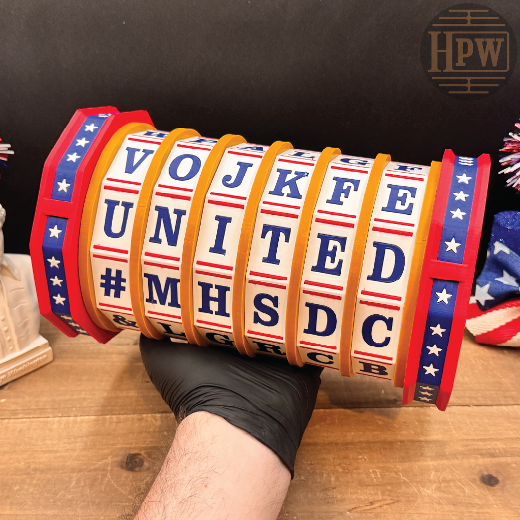 A novelty alphabet wheel sign held by a gloved hand on a wooden table, featuring red, white, and blue patriotic borders with orange lettering bands.