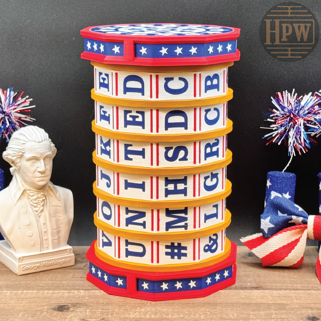 A patriotic-themed wooden cylindrical box with stacked letter compartments spelling out 'UNITED STATES' and '#VOTEDMIGHTY'. The box features a red, white, and blue top and base with white stars, and is styled on a wooden surface with a George Washington bust, a small American flag, and decorative pom-poms against a dark background.