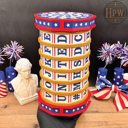 A patriotic-themed cylindrical side table or stool with a red, white, and blue striped top and base, yellow bands, and white panels spelling out 'UNITED STATES' and 'VOTE'. The item is held by a black-clad hand on a wooden surface, with patriotic decorations in the background.