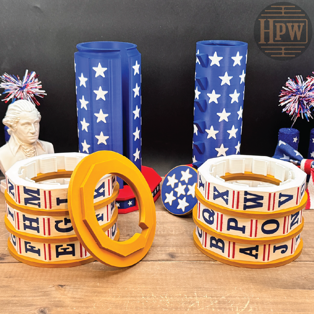 Alphabet ring necklaces displayed on a wooden surface, featuring white rings with red and blue stripes and black letters, alongside matching blue and white star-patterned cylindrical holders.