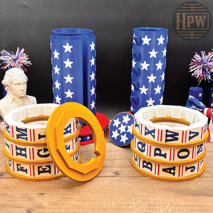 Alphabet ring necklaces displayed on a wooden surface, featuring white rings with red and blue stripes and black letters, alongside matching blue and white star-patterned cylindrical holders.