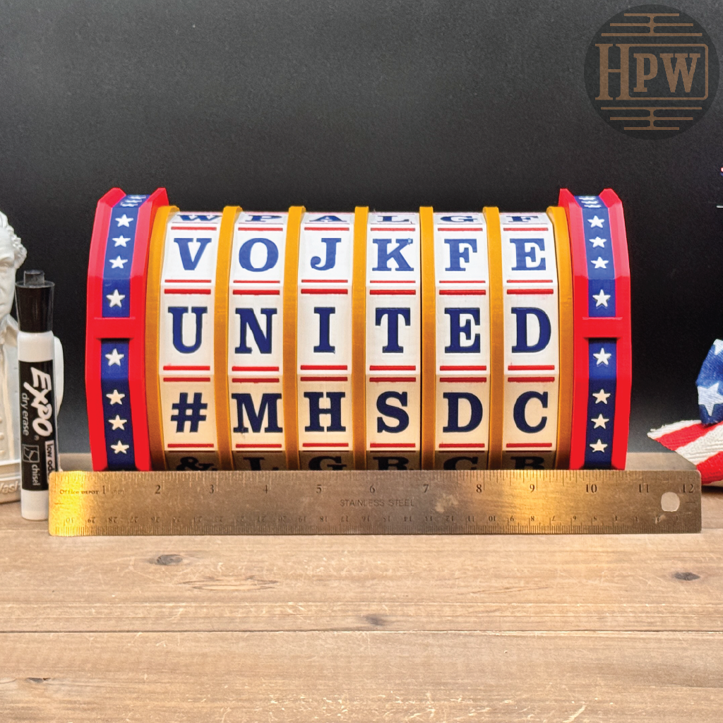 A novelty flip sign displaying patriotic American colors and the text 'VOJ KFE UNITED #MHSDC' with a gold-colored metal ruler placed in front for scale, set on a wooden tabletop against a dark background.