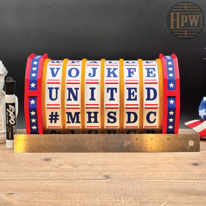 A novelty flip sign displaying patriotic American colors and the text 'VOJ KFE UNITED #MHSDC' with a gold-colored metal ruler placed in front for scale, set on a wooden tabletop against a dark background.