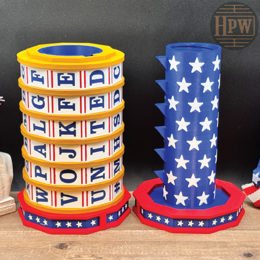 Two patriotic-themed cylindrical party favors or containers on a wooden surface against a dark background. One is stacked with white bands featuring blue and red text, bordered in yellow, and sits on a red, white, and blue base.