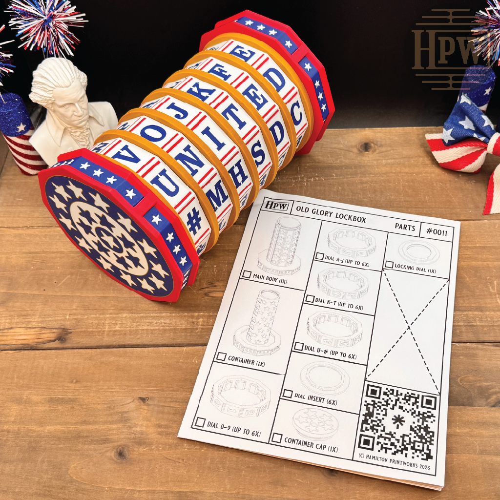 A patriotic-themed cylindrical lockbox with red, white, and blue patriotic stripes and stars, displayed on a wooden table next to an instruction sheet. The lockbox is partially open, revealing the interior, with decorative American flags and a bust in the background.