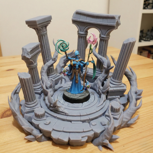 A painted fantasy tabletop miniature figurine of a blue-robed female character standing on a circular stone platform, surrounded by gray ruined columns, twisted tree branches, and a pink flower, photographed on a wooden tabletop.