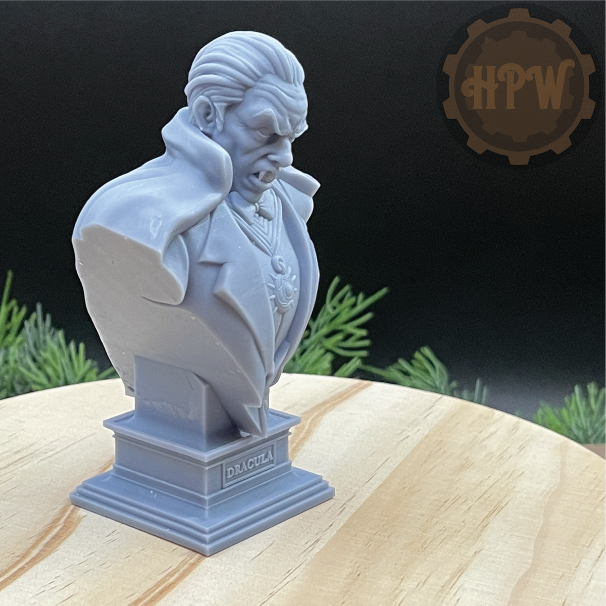 A light gray 3D-printed resin bust of a vampire character on a pedestal base, displayed on a light wood surface with a dark background and green foliage props.