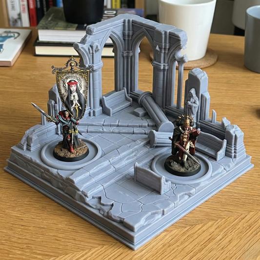 A gray tabletop miniature terrain piece depicting ruined gothic arches and columns, with two painted fantasy miniatures standing on circular platforms within the set, photographed on a wooden desk.