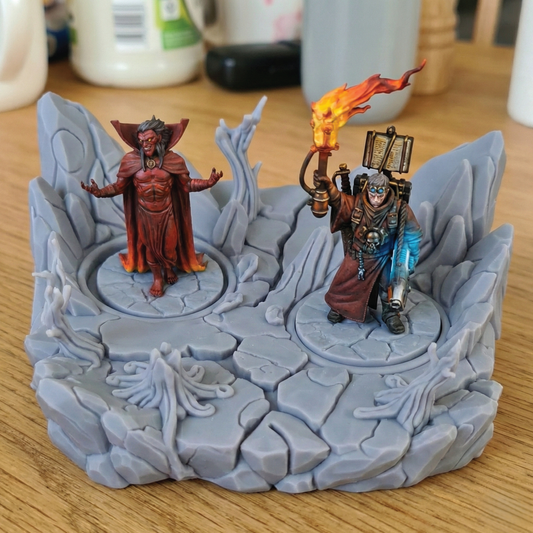 A miniature tabletop gaming terrain piece featuring a gray rocky platform with two fantasy character figures, one in a red robe and one in a brown robe holding a flaming torch, displayed on a wooden tabletop.