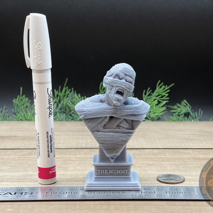 A small gray mummy bust sculpture displayed on a wooden surface with a ruler, coin, and marker for scale, against a dark backdrop with blurred greenery.