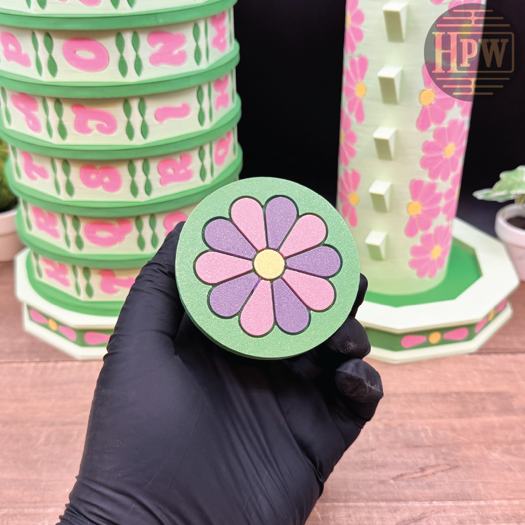 Hand holding a floral coaster with stacked coasters and a pink cup in the background.