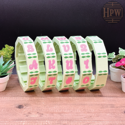 Set of letter-shaped clips with pink letters on a wooden surface with plants in the background.