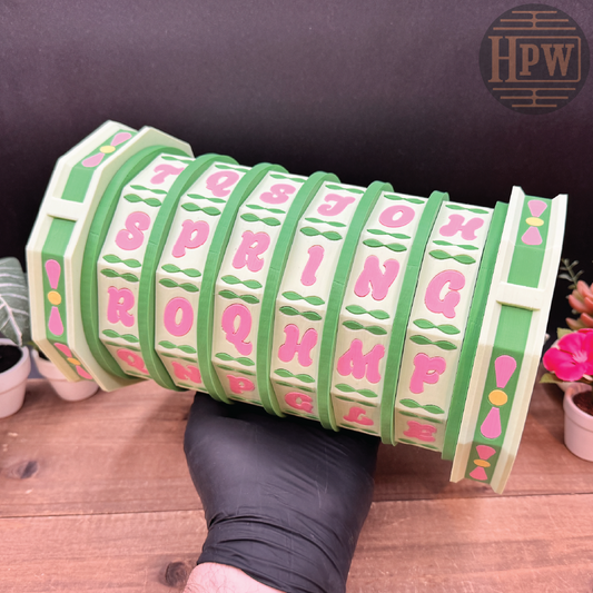 Green and pink lettered cylindrical object held by a gloved hand on a wooden surface with plants in the background.