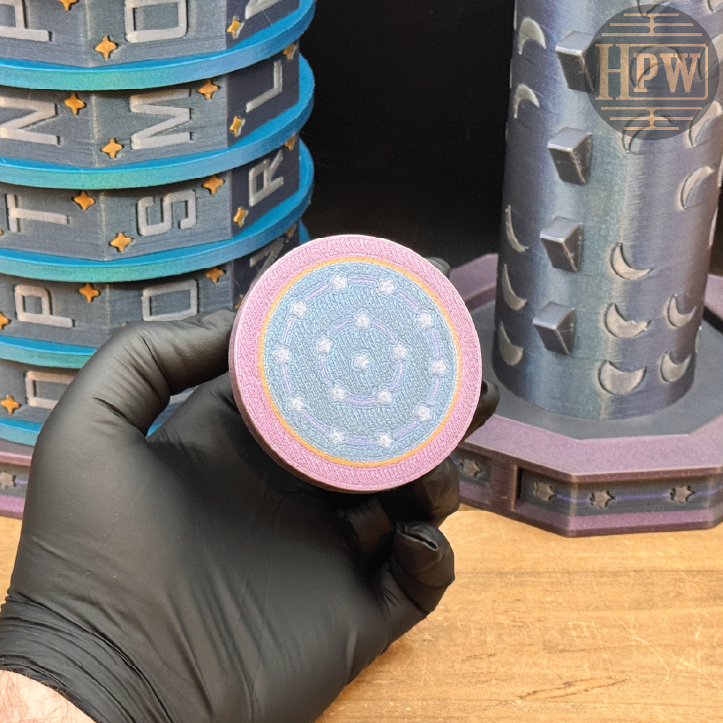 A hand wearing a black glove holds a round, patterned item with a pink outer ring and a blue center featuring white celestial motifs. In the background, stacked blue and gray cylindrical items and a patterned gray column are visible on a wooden surface.