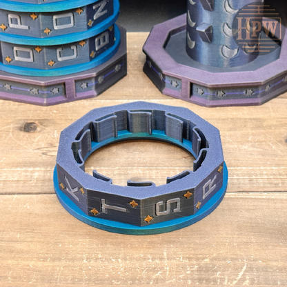 A 3D-printed miniature accessory, specifically an octagonal ring collar with blue and purple layers and gold symbols, displayed on a wooden tabletop alongside other similar miniature parts.