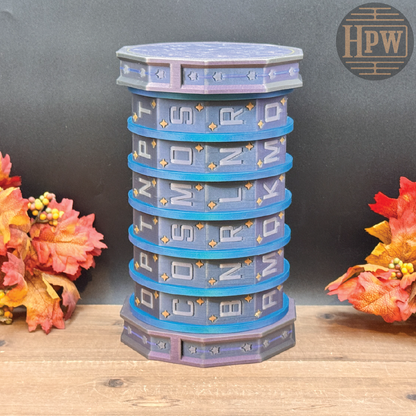A decorative cylindrical stack of hexagonal boxes in blue, purple, and gray, each labeled with a letter, displayed on a wooden surface with autumn leaves on both sides.