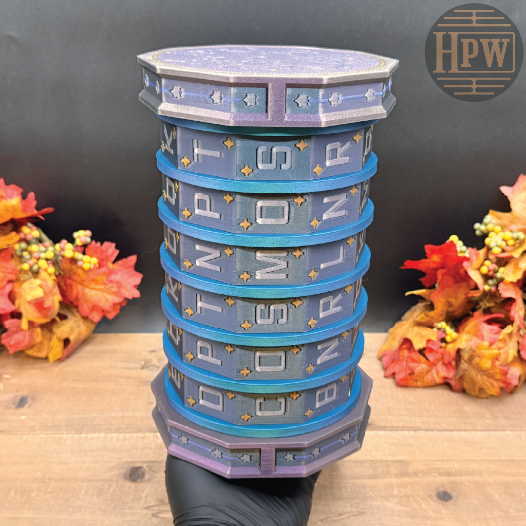 A decorative cylindrical stack of six stacked round boxes or containers in blue, teal, and lavender with gold accents and white lettering, held by a black gloved hand on a wooden surface with autumn leaves on both sides. An HPW logo is visible in the top right corner.