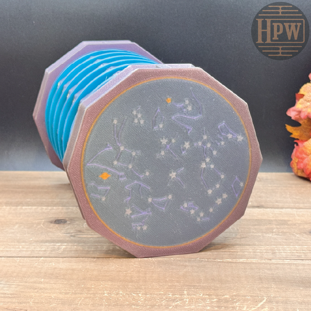 A cylindrical octagonal speaker or diffuser with a gray front face printed with purple constellations and small orange stars, surrounded by a brown rim and blue inner panels, shown on a wooden surface against a dark background.