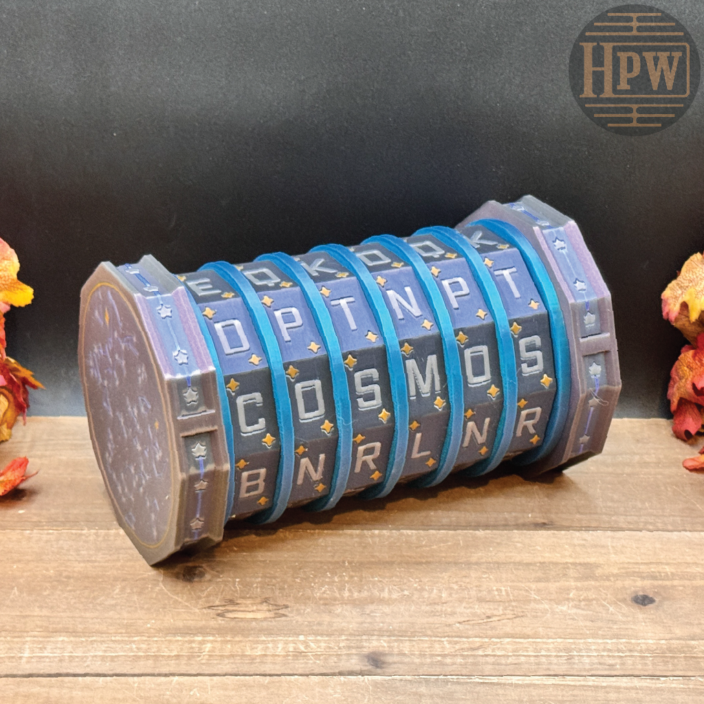 A cylindrical, multi-ring mechanical puzzle box with blue and gray segments displaying letters, resting on a wooden surface against a dark background with decorative autumn leaves.