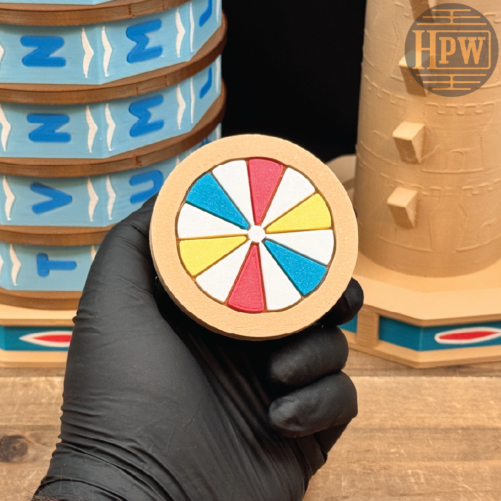 A hand wearing a black glove holds a round wooden token or coin featuring a colorful wheel design with red, yellow, blue, and white segments. In the background, wooden toy structures and stacked blue-and-beige pieces are visible on a wooden surface.