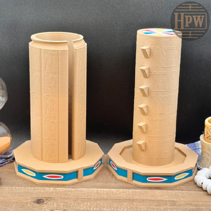 Two tan cylindrical decorative objects with carved-looking vertical patterns and multicolor band details at the base, displayed on a wooden surface against a dark backdrop. A circular HPW mark appears in the top right.