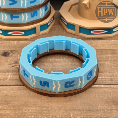 A light blue 3D-printed ring-shaped object with raised letter segments and a brown circular base, displayed on a wooden surface with similar objects blurred in the background. A circular HPW logo watermark is visible in the top right corner.