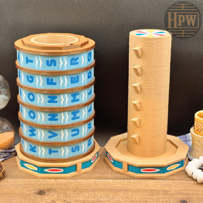 Two wooden toy game towers displayed on a wooden tabletop against a dark backdrop. The left tower has a blue and tan striped pattern with alphabet letters, and the right tower is a natural wood cylinder with carved details and small colored accents at the top and base.