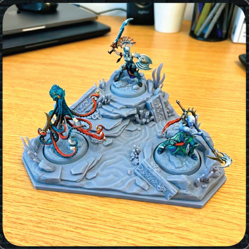 A painted tabletop miniature terrain piece featuring three fantasy figures on a gray rocky base, photographed on a wooden desk with office items blurred in the background.
