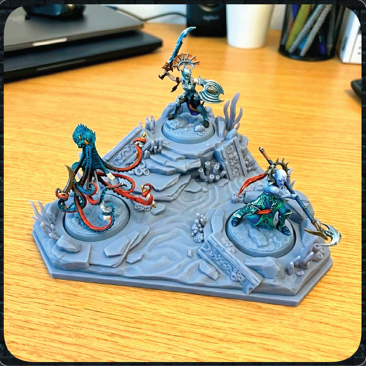 A painted tabletop miniature terrain piece featuring three fantasy figures on a gray rocky base, photographed on a wooden desk with office items blurred in the background.