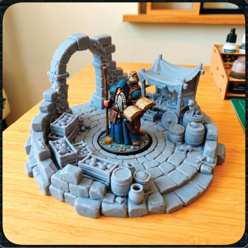 A painted tabletop miniature scene on a round stone platform, featuring a robed wizard with a staff and an open book, a small stall with barrels and crates, and a ruined archway backdrop, displayed on a wooden tabletop.