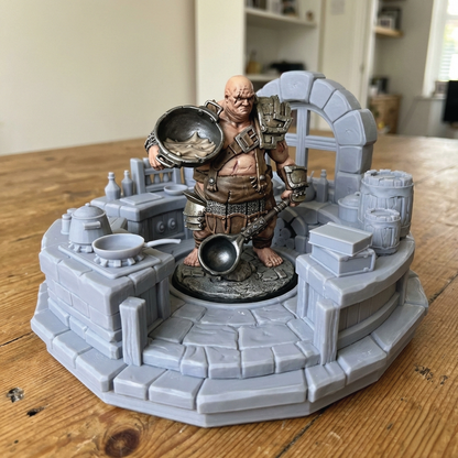 A painted tabletop miniature figurine of a muscular barefoot warrior holding a large metal cauldron, posed on a gray stone platform with barrels, crates, and architectural details, set on a wooden table.